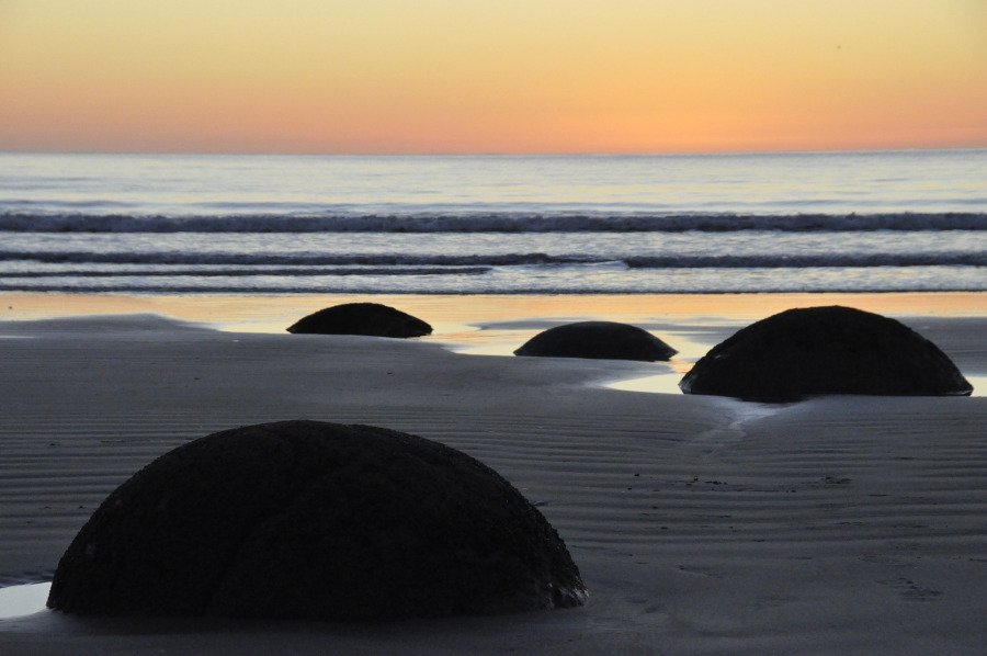 Strand mit vier runden Felsen im Vordergrund, Meer mit leichten Wellen im Hintergrund, Sonnenuntergandsstimmung
